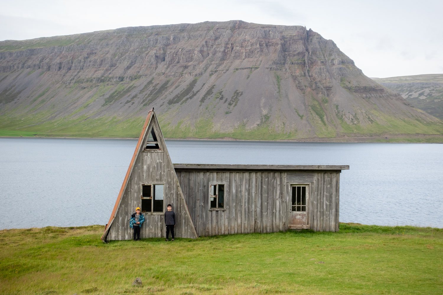 Chris Burkard's 2 sons stand in front of a wooden cabin with a fjord and mountain backdrop, at Fossfjörður in the Westfjords