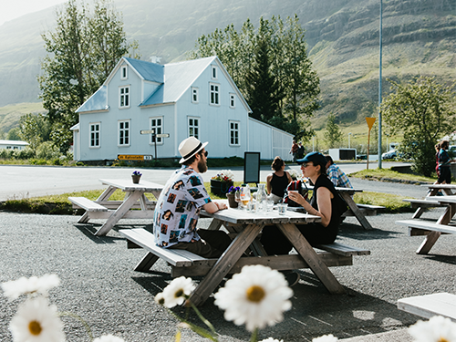 Zwei Personen sitzen auf einer Picknickbank vor einem Lokal in Seyðisfjörður und Gänseblümchen blühen im Vordergrund 