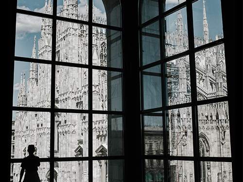 A person stands in silhouette against a very large window, overlooking the Duomo di Milano 