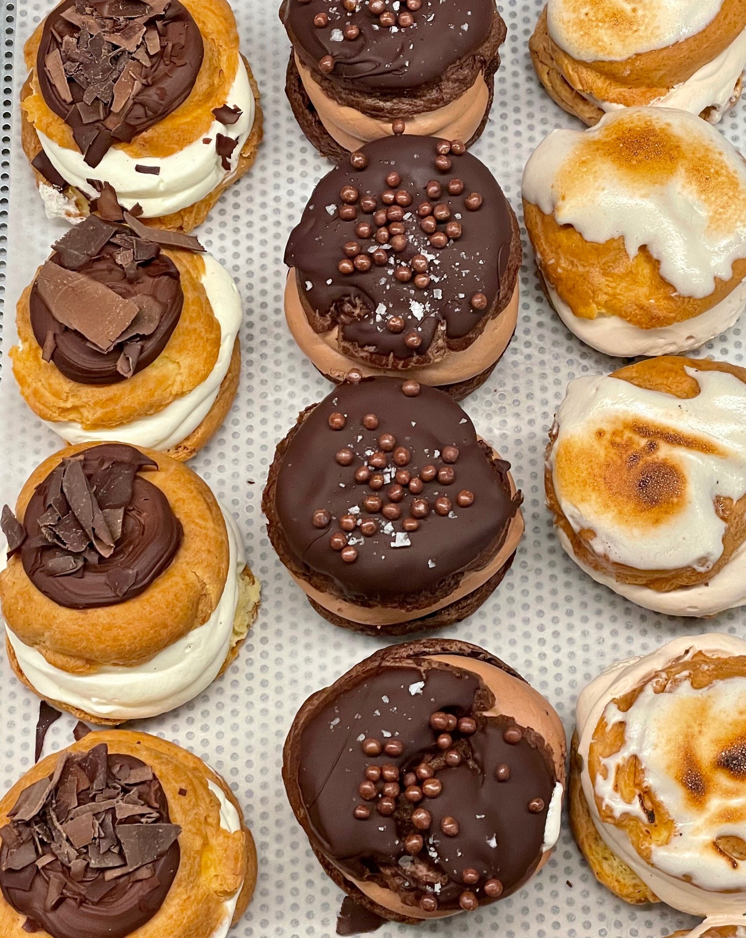 An overhead image of nine cream buns in a Reykjavík bakery. There are three different flavours: regular topped with chocolate sprinkles, regular topped with lemon meringue, and chocolate buns topped with chocolate icing.