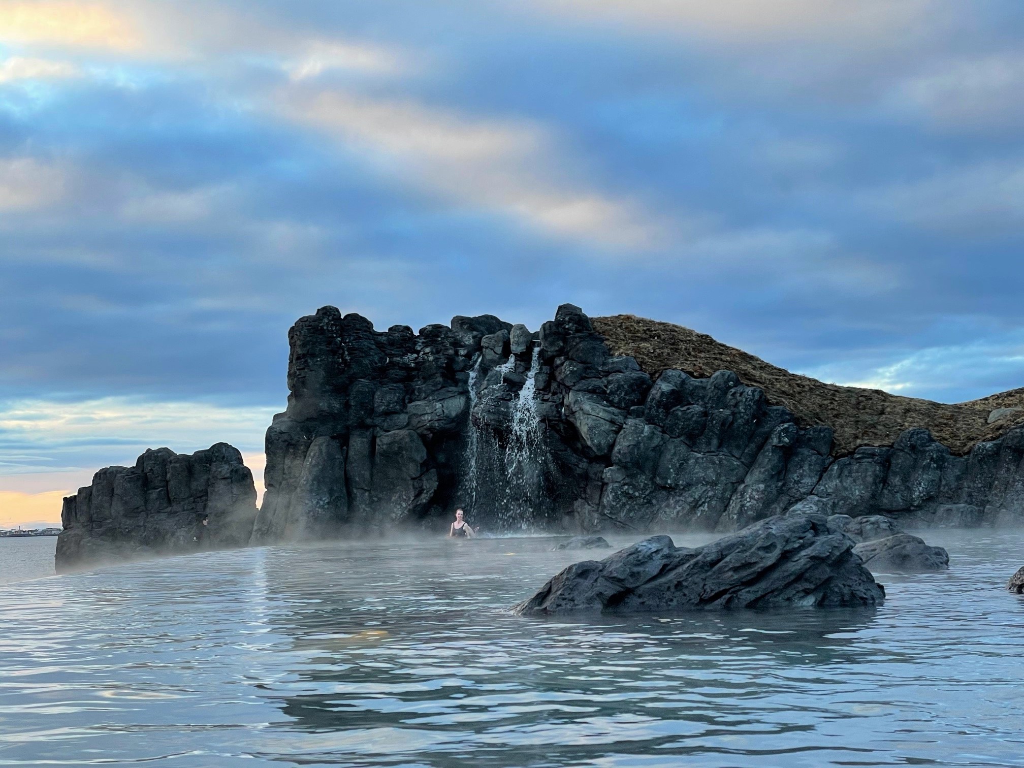 A person bathing in the Sky Lagoon in Reykjavik, pictured with water pouring over the black basalt cliffs onto themselves and into the steaming pool