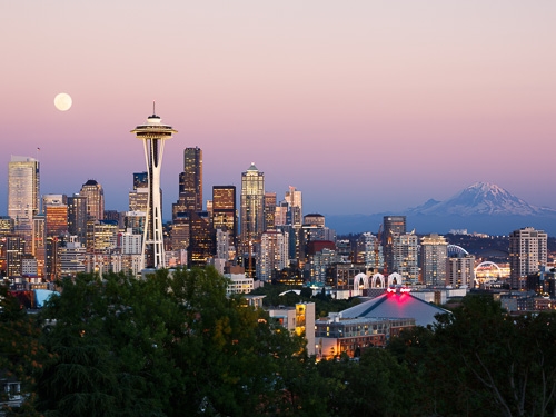 A skyline scene of Seattle as pictured at sunset, with a bright moon and mountains in view 