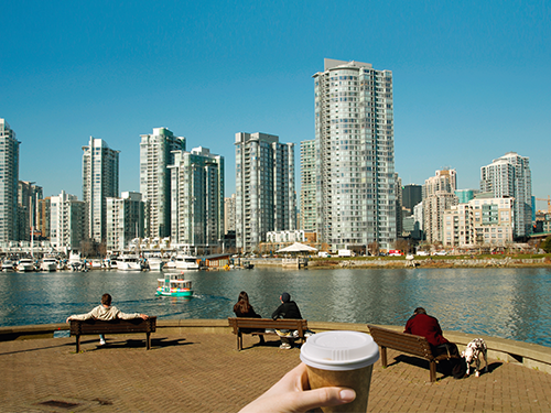 A hand holds a coffee cup up to the frame. Behind the cup there are people sitting on benches facing a cityscape of Vancouver 
