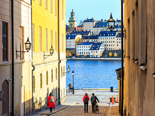 A view to the harbour in Stockholm as seen from between two buildings. A couple walks down the street in the foreground
