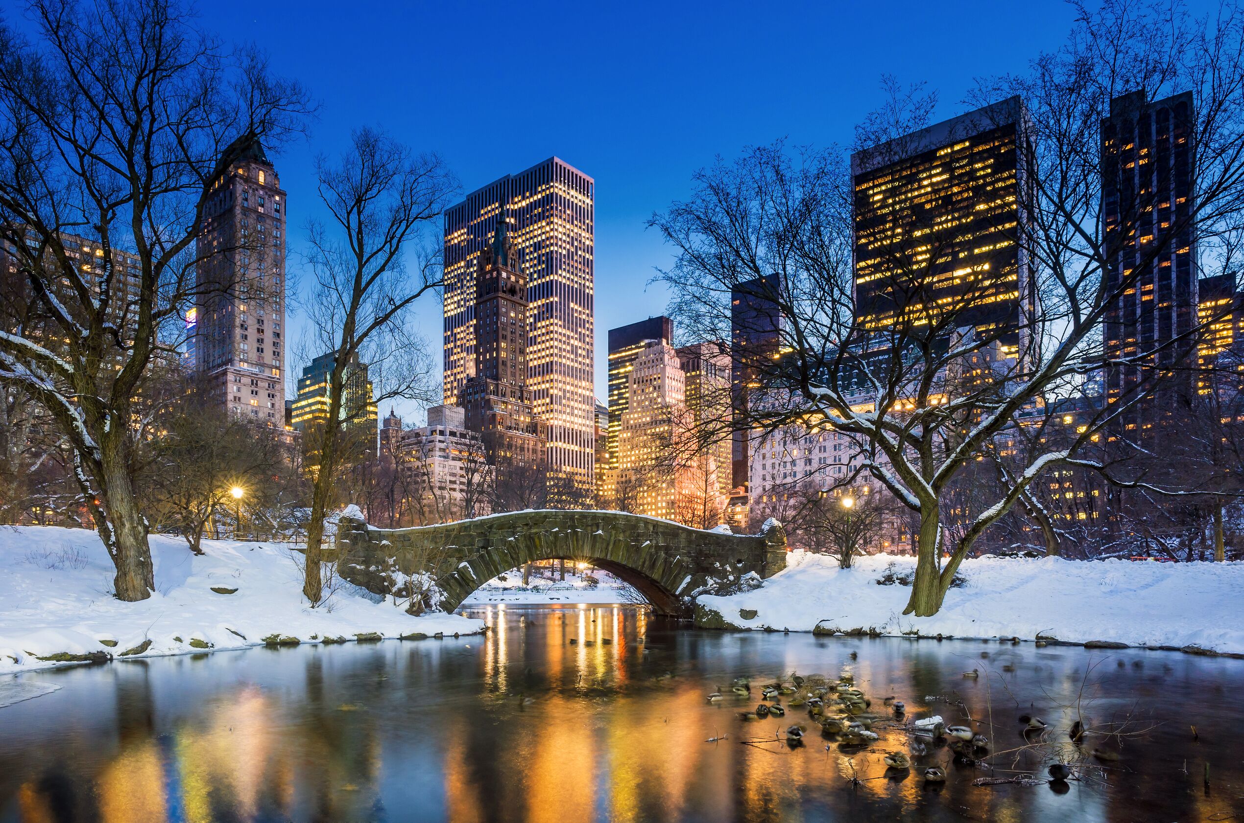A stone bridge in Central Park at dusk, with snow opn the ground and the lights of the cuty building's reflected in the water