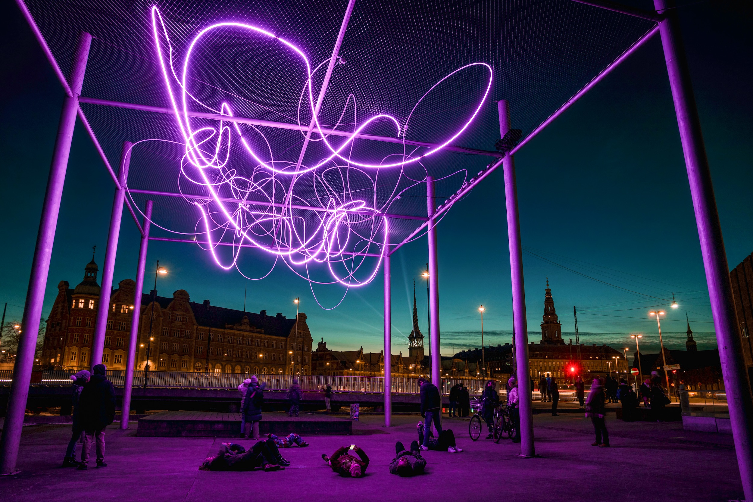 People lying down and standing outdoors beneath a light art installation. The strings of light are thin, purple and swirly, supported by tall metal columns and wire mesh.