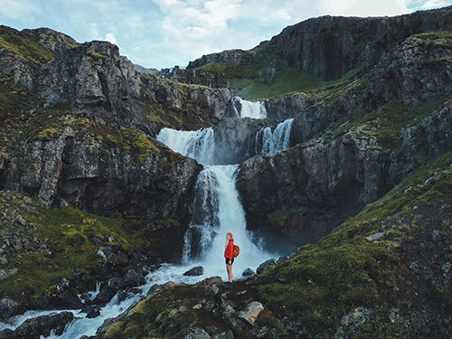 Een vrouw in een rode jas staat voor de prachtige Klifbrekkufossar waterval in Oost-IJsland 