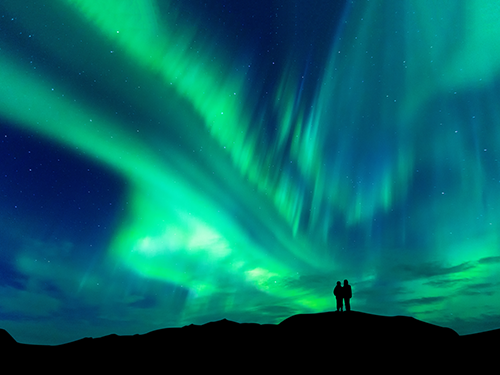 Two people pictured as silhouettes in the distance while the Northern Lights light up the sky overhead