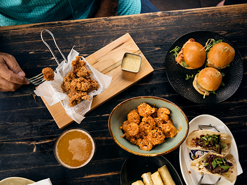 A man’s hand holding a fork is pictured with fried chicken on it, positioned next to fried tempura prawn, burgers, coffee and tacos 