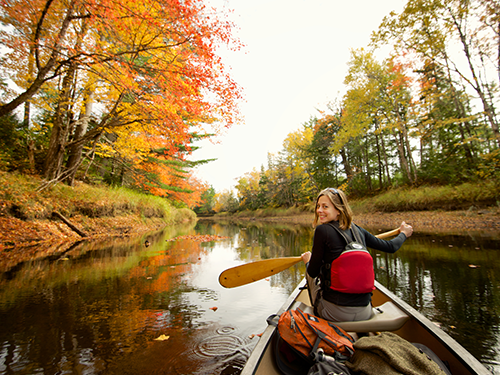 A woman looks back to the camera as she canoes down a river in America, with the trees lit up in autumnal colours 
