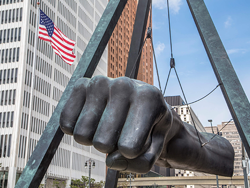 The First sculpture pictured from the front-on, set against a blue sky day, with an American flag positioned to the left hand side of the picture