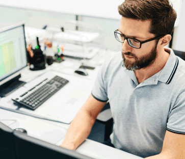 35-year-old man with glasses wearing a polo shirt, sitting in a bright modern office desk, typing on a desktop computer.