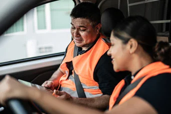 Two drivers in a semi-truck cab wearing high visibility vest looking toward at a tablet