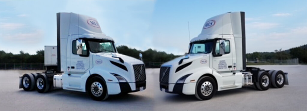 Two MLS tractors parked nose-to-nose in a facility parking lot with trees in the background.