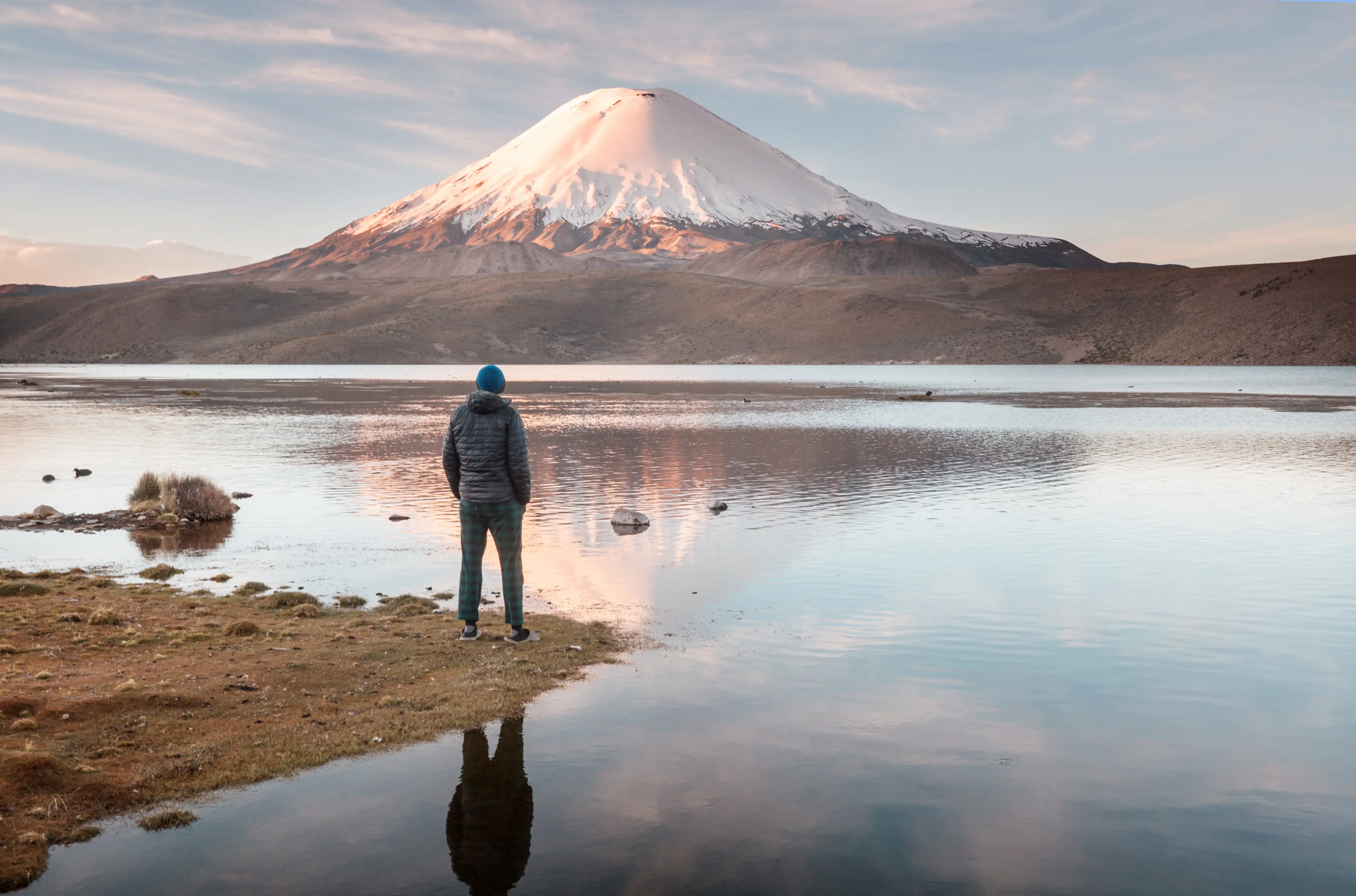Vulcão nevado refletido em lago no altiplano do Atacama, com viajante contemplando a paisagem. Viaje com pontos na Livelo.