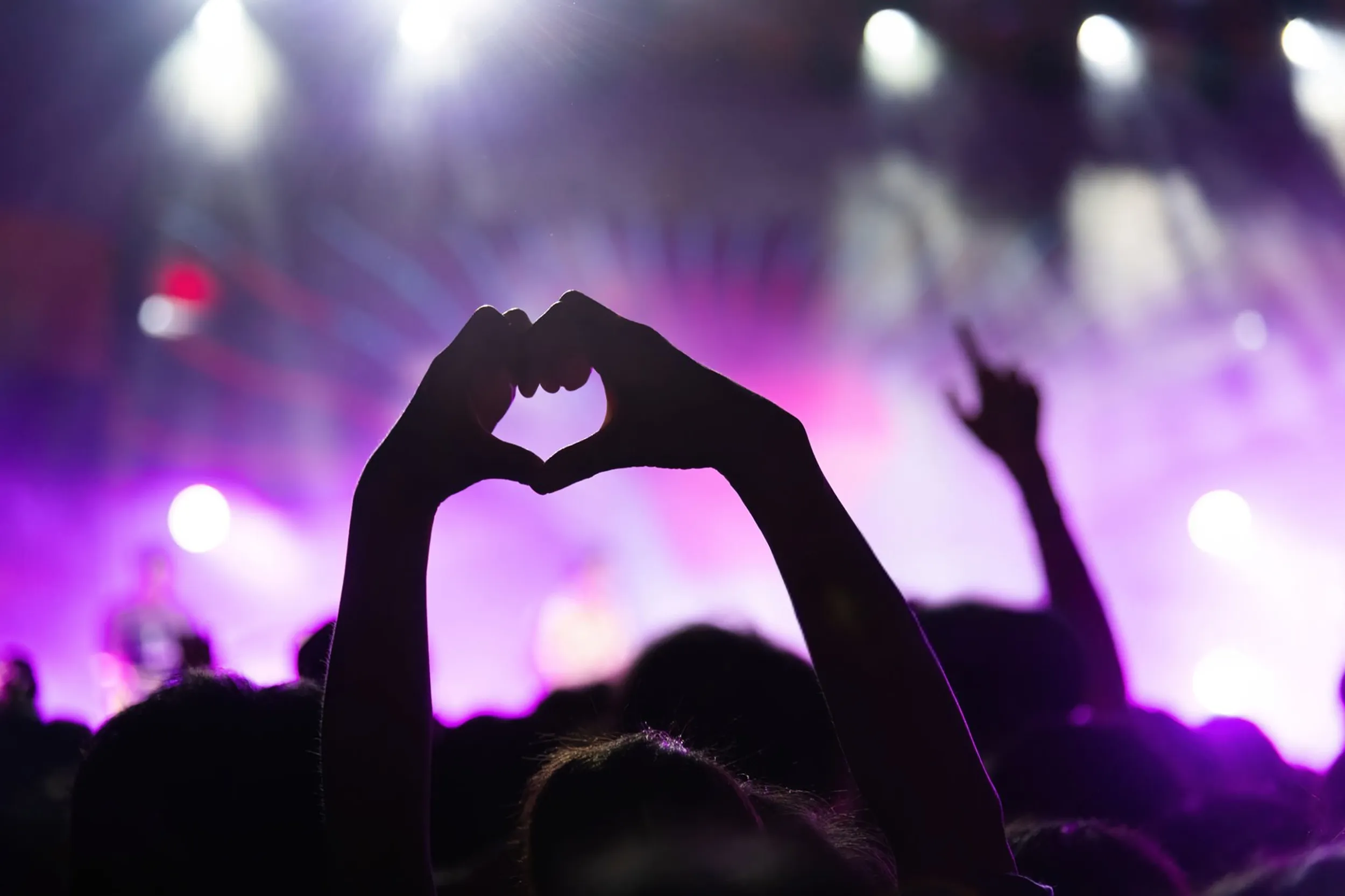 Silhueta de mãos formando um coração durante Lollapalooza em Chicago, com palco iluminado em tons de rosa e roxo. Viva momentos especiais com pontos Livelo.