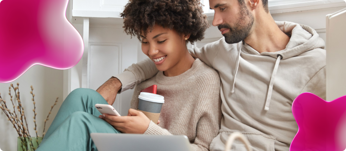 Um casal olha para o celular na mão da mulher enquanto estão sorridentes e se abraçando sentados em um sofá.