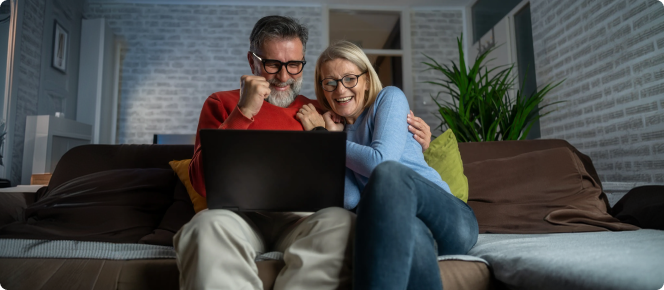 Casal sentado no sofá comemorando alegremente ao ver o conteúdo na tela de um notebook.