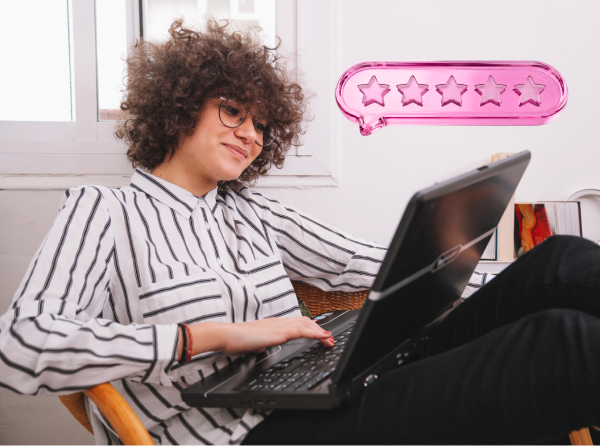 Mulher olha sorridente para o notebook que está no seu colo, ao seu lado existe o desenho de um balão em rosa com 5 estrelas.