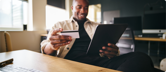 Homem sorridente olha para o celular em uma mão e um cartão de crédito na outra.