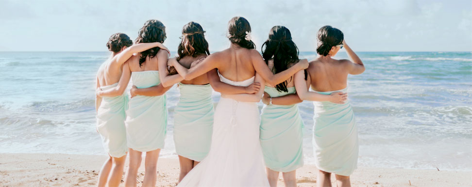 A bride and her bridesmaids look out into the ocean from a beach.