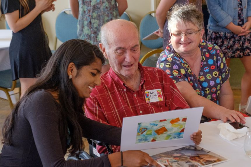 Student and an elderly man and woman sit at a table and look at artwork.
