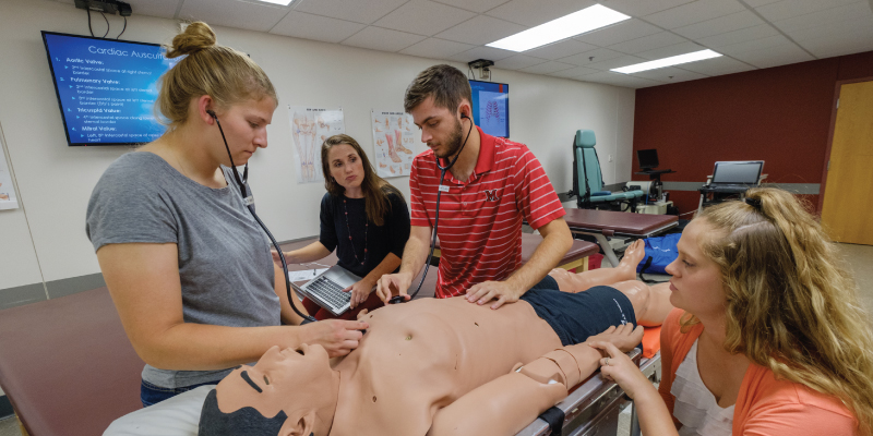 3 students and their professor using a stethoscope on a mannequin.
