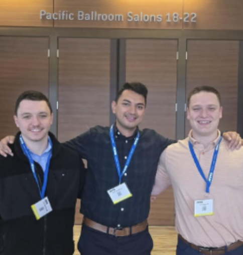 Three men stand in front of a sign reading "Pacific Ballroom Salons," smiling and posing for the photo.