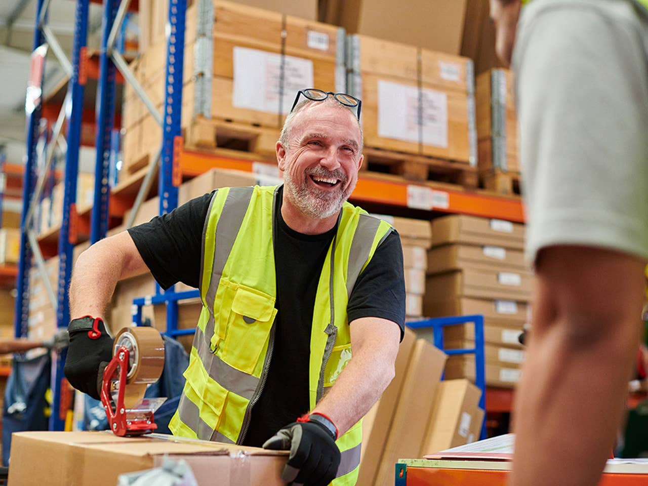 A smiling man stands in a warehouse surrounded by stacked boxes.