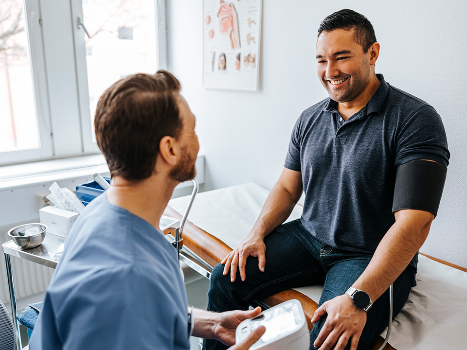 Smiling young man wearing a grey polo shirt sits on an exam room table and speaks with a Corewell Health Occupational Health specialist