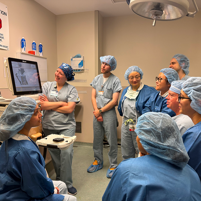 Medical trainees in surgical scrubs gather around a monitor during instruction in an operating room.