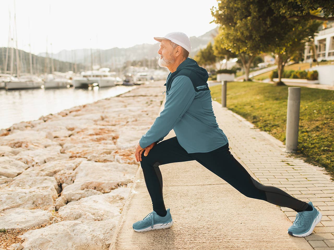Older man wearing black jogging pants and a blue jogging jacket does a lunge stretch while outside