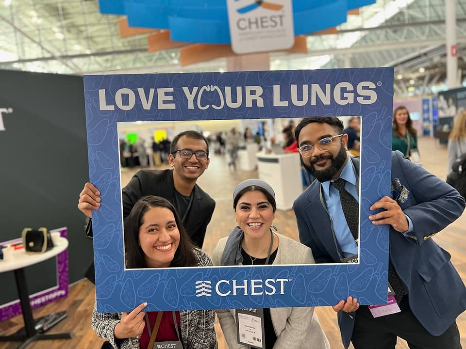 Residents pose at a medical conference holding a “Love Your Lungs” CHEST-branded photo frame on an exhibit hall floor, highlighting participation in a national pulmonary medicine event.