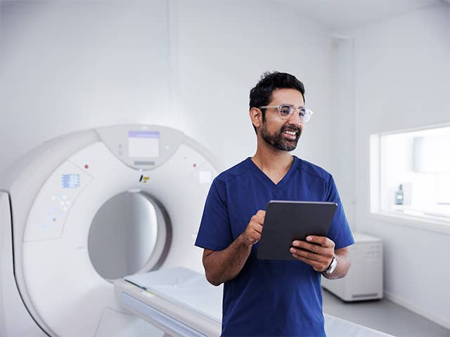 Man with glasses and a beard and wearing blue scrubs stands in front of an MRI machine