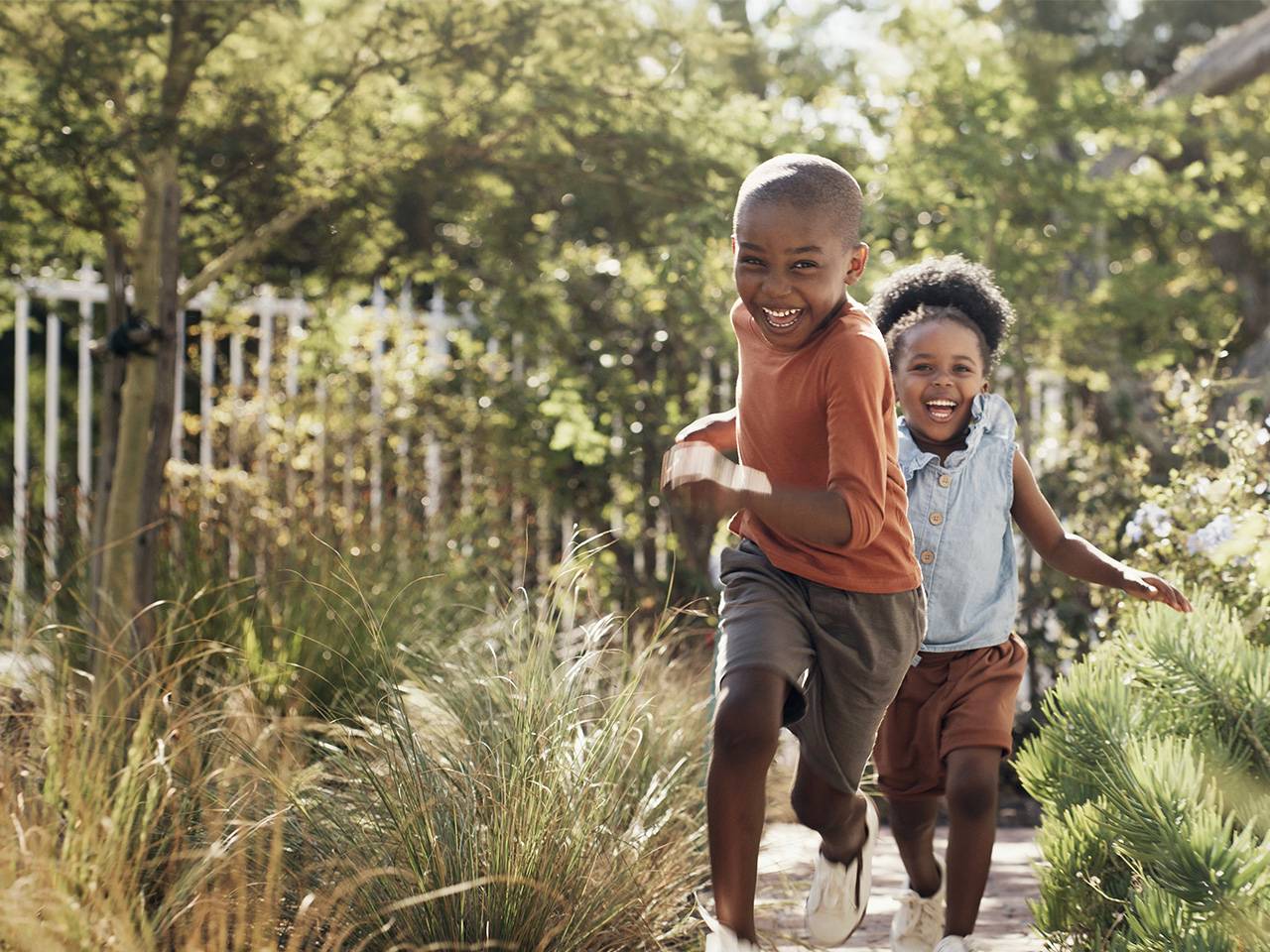 Smiling young Black boy and his smiling sister run together through tall wild grass in the sunshine outdoors