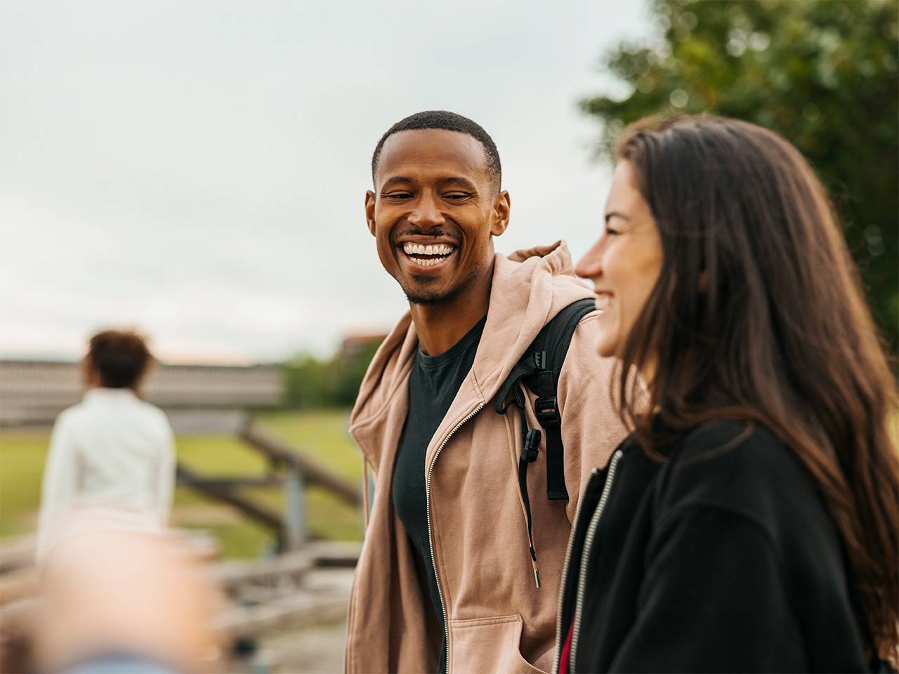 Young Black man wearing a light brown jacket smiles and laughs while talking with a woman outdoors