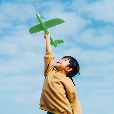 Boy with black hair and wearing a yellow shirt holts a green airplane toy into the sky