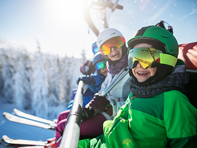 Three skiers in colorful gear seated on a ski lift, surrounded by snowy mountains and clear blue sky.