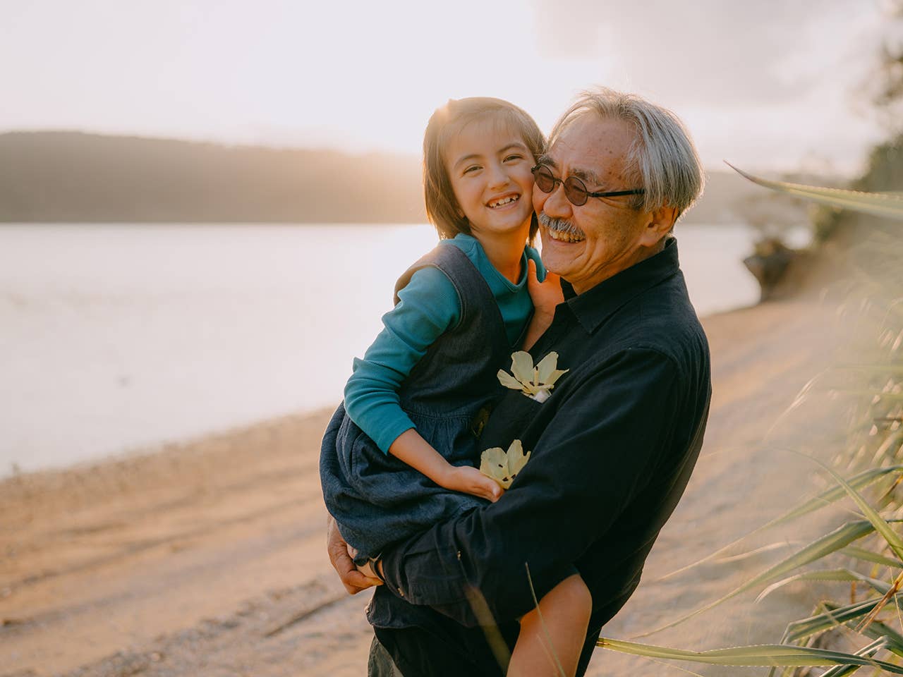 Elderly Asian man lifts up his granddaughter as they stand on a beach