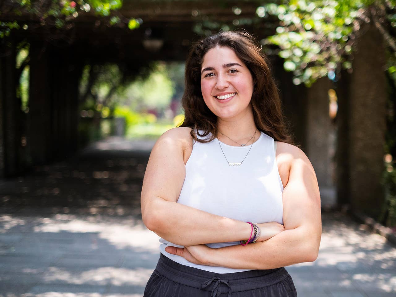 Smiling young white woman wearing short-sleeved workout clothing stands in the shade outdoors