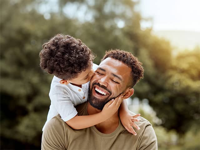 Smiling young Black man carries his son on his back as they play outdoors