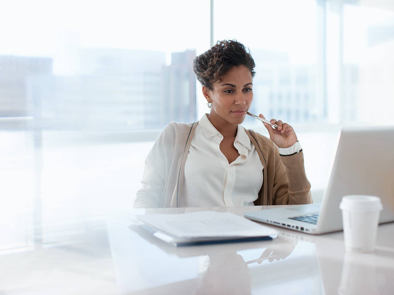 Black woman in a white blouse sits at a desk and looks at her laptop