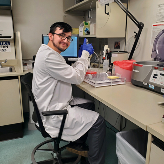 Male student in white lab coat sitting at a bench testing something out