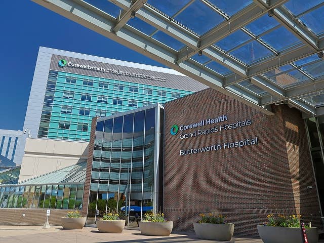 Exterior view of Corewell Health Grand Rapids Hospitals Butterworth Hospital entrance with glass canopy, brick wall signage, and modern multi-story building in the background under a blue sky.