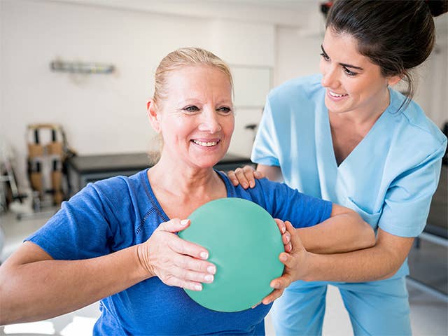 A woman holds an orange ball while a nurse assists her during an Occupational Therapy session in Physical Rehabilitation.