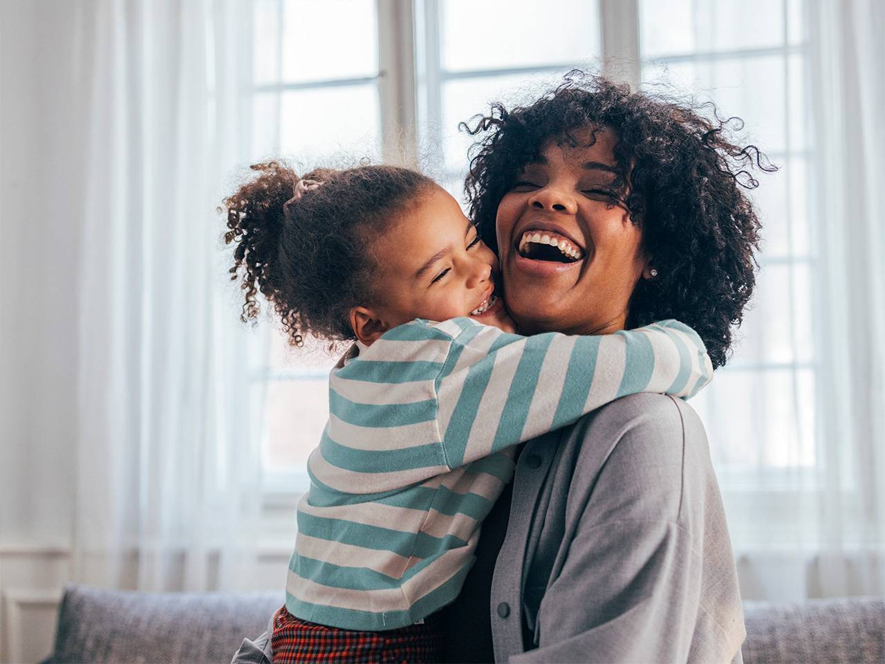 Smiling mother with curly black hair lifts up and holds her smiling daughter, who is giving her a hug