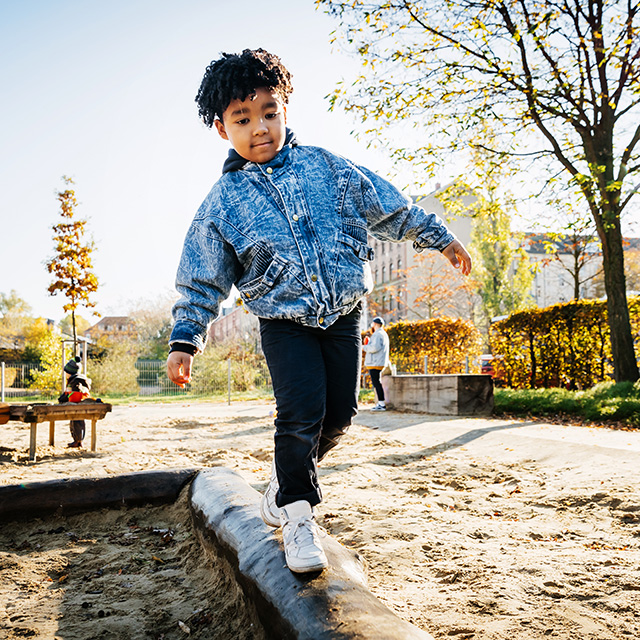 A Black boy wearing a denim jacket walks along a balance beam in a playground outdoors