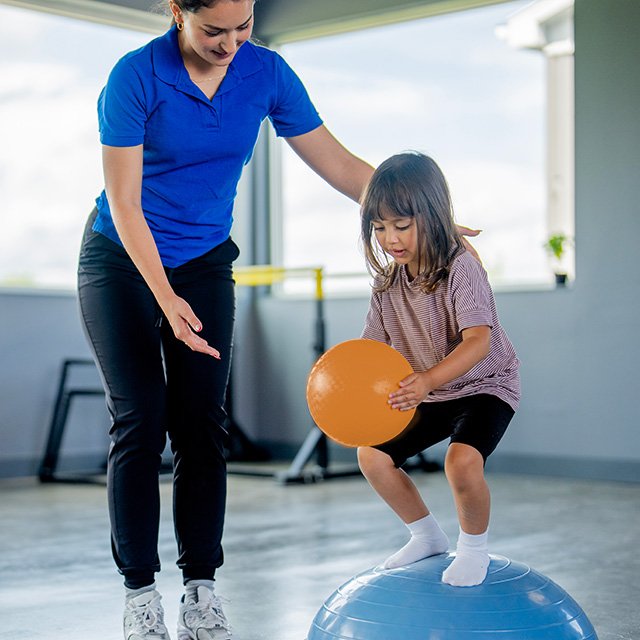 A Corewell Health pediatric physical therapist helps a young girl balance on therapy equipment in an Helen DeVos Children's Hospital clinic