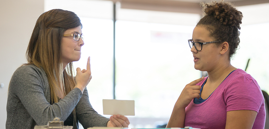 Two women at a desk in an office, one delivering speech therapy while the other listens attentively