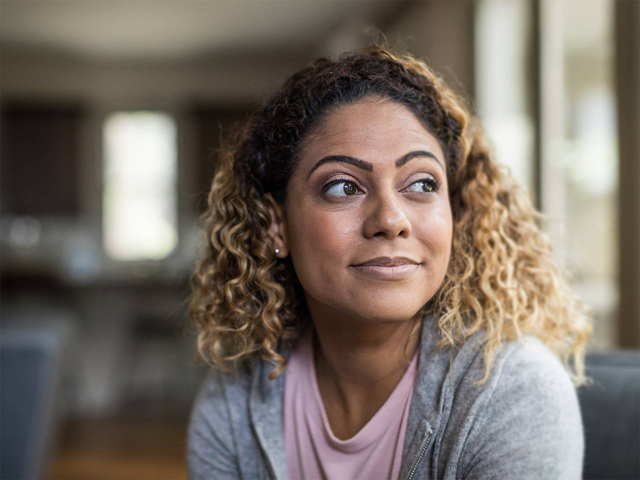 Black woman wearing a pink blouse and grey sweater sits in a sunlit kitchen